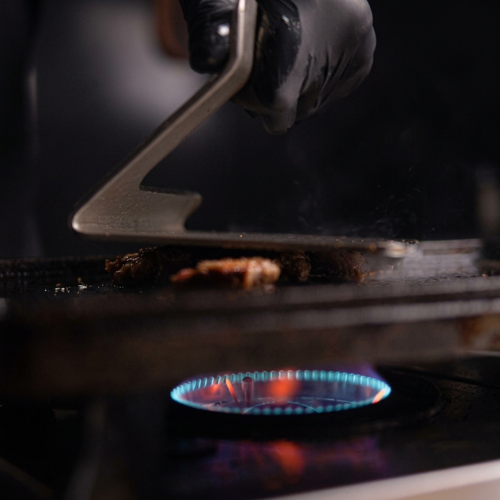 A gloved hand using a metal grill press to smash a mound of pulled pork flat onto a hot cast iron griddle with blue flames visible underneath.