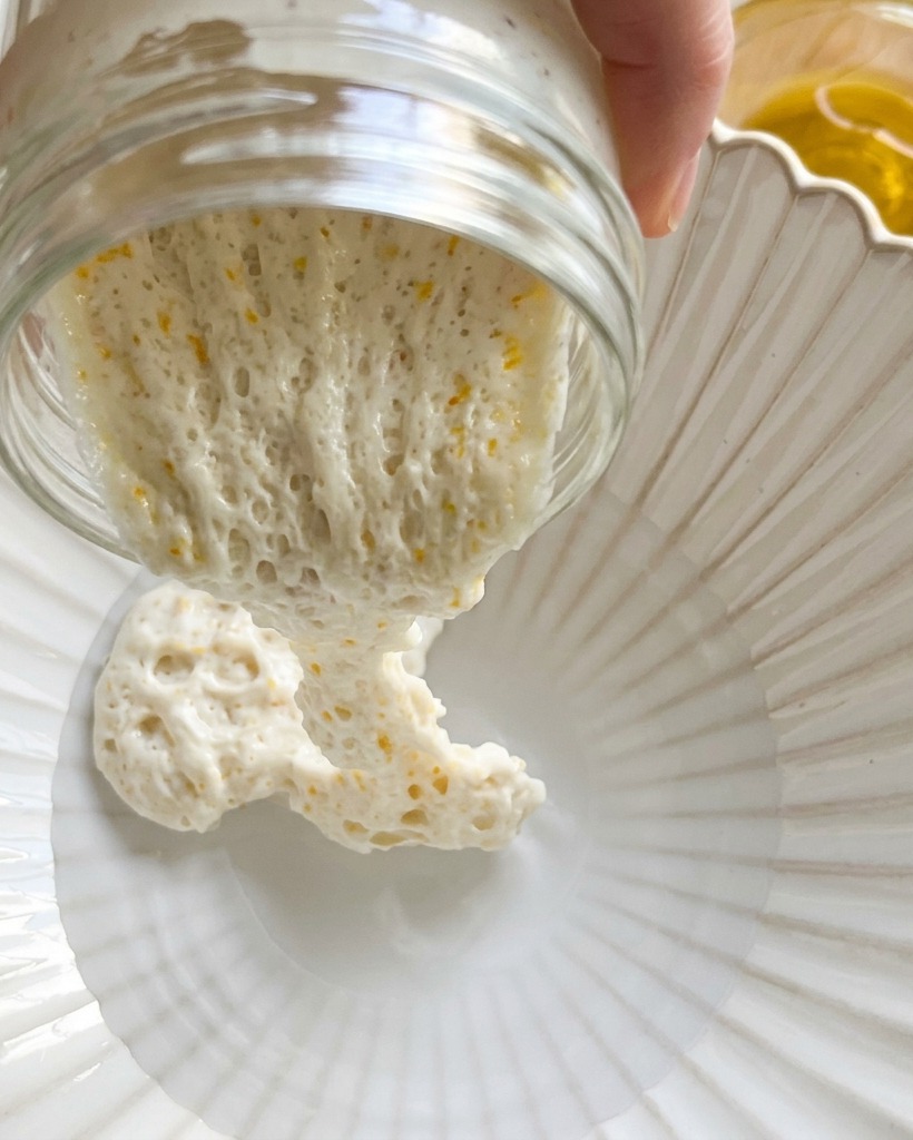 Dry flour being poured into a bowl containing water and levain for the initial dough mix.