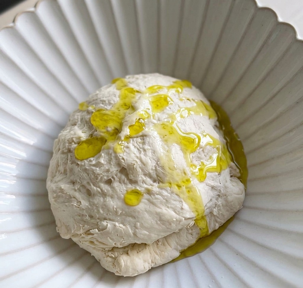 Olive oil being poured over a ball of dough that is sitting in a white bowl.