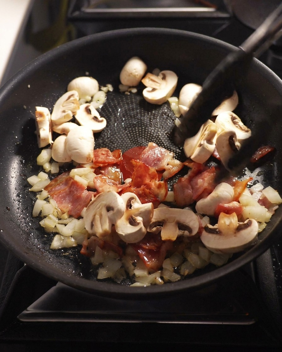 Sliced button mushrooms being stir-fried in a pan with bacon, onions, and garlic.