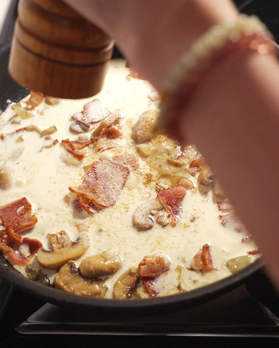 A hand using a wooden pepper grinder to crack fresh black pepper over a pan of simmering creamy sauce with bacon and sliced mushrooms.