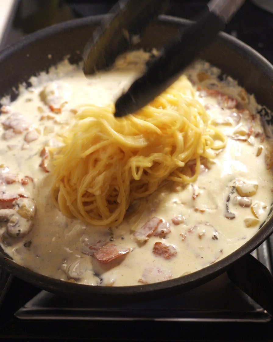 Black kitchen tongs dropping a portion of freshly cooked spaghetti into a pan filled with a white cream sauce, bacon pieces, and mushrooms.