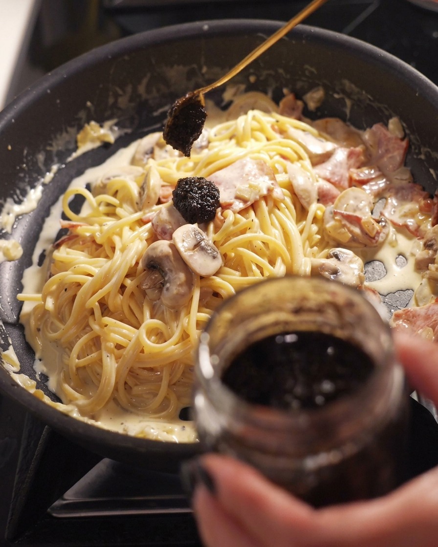 A spoon dropping a dark dollop of black truffle paste from a jar onto a pan of creamy spaghetti mixed with bacon and mushrooms.