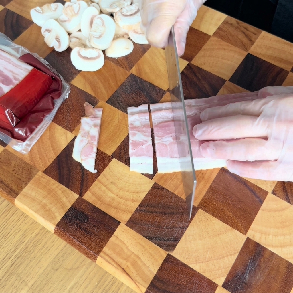 Gloved hands using a kitchen knife to cut raw bacon strips into smaller rectangular pieces on a checkered wooden cutting board.