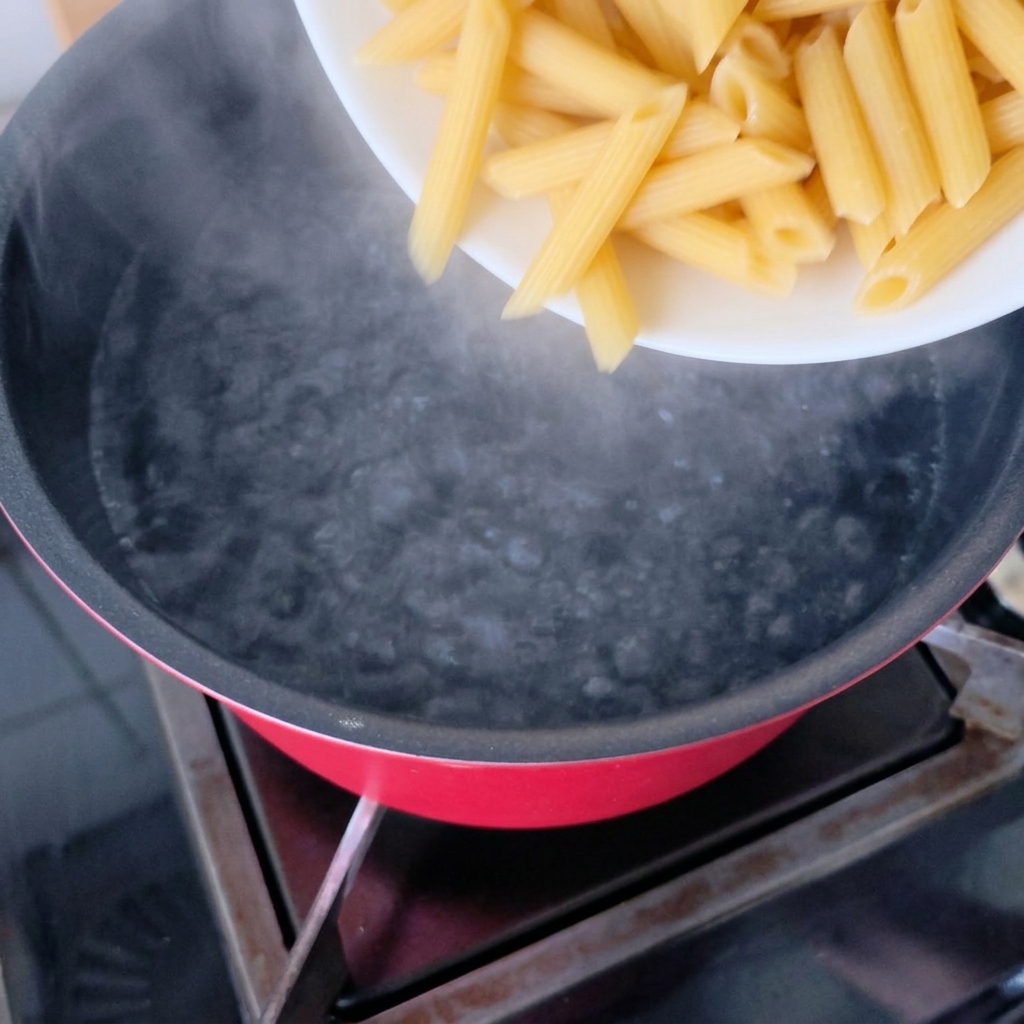 Dry penne pasta being poured into a pot of boiling water.