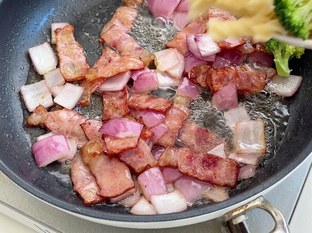 Cooked fusilli pasta and bright green broccoli florets being poured into a pan with sizzling bacon and red onions.