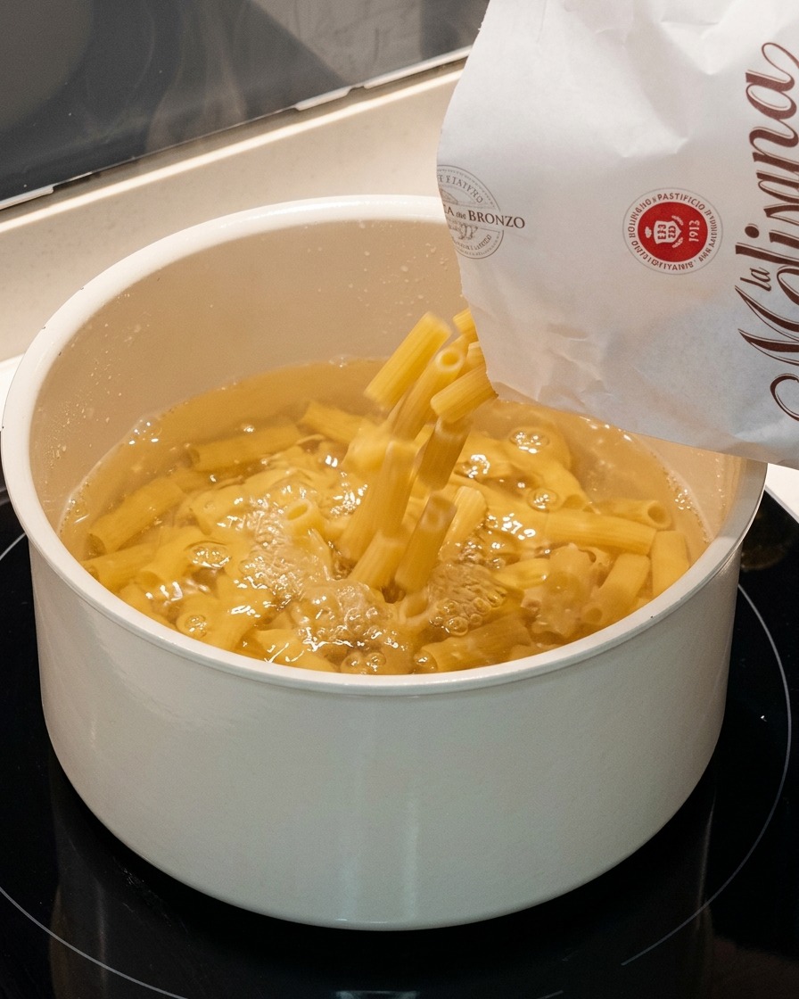 Dry tube pasta being submerged into a large pot of bubbling water.