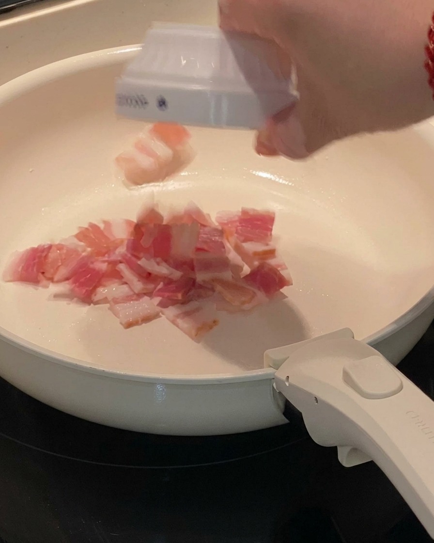 Small pieces of bacon sizzling in a flat pan, releasing their fat.