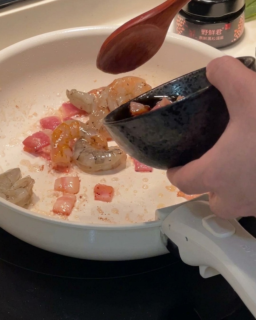 Shrimp pieces being added to a pan containing rendered bacon.
