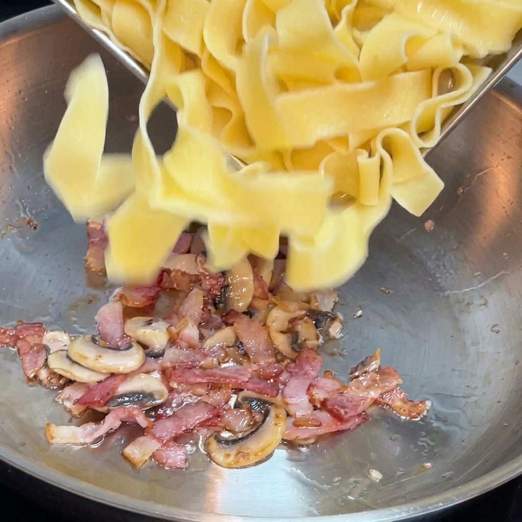 Freshly cooked wide pasta noodles being tossed into a skillet filled with cooked bacon bits and sliced mushrooms.