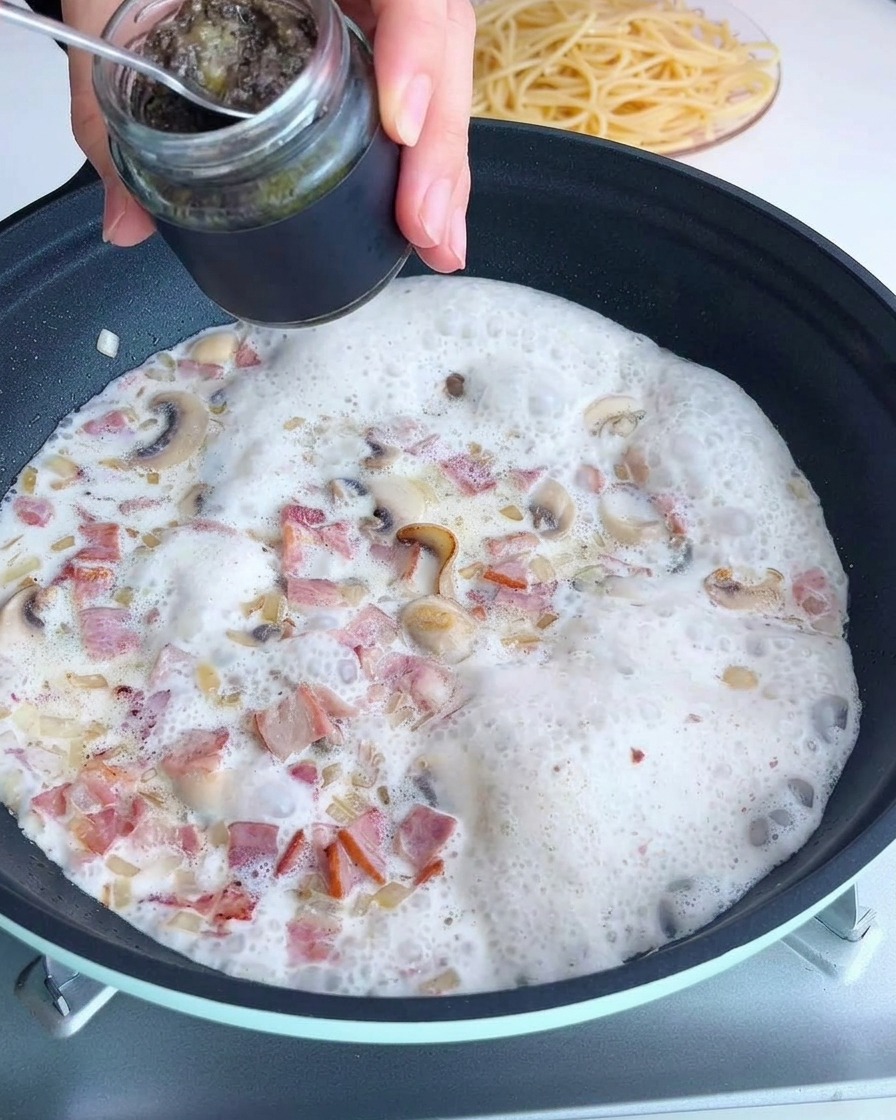 A spoonful of black truffle sauce being added to a pan with a creamy mushroom mixture.