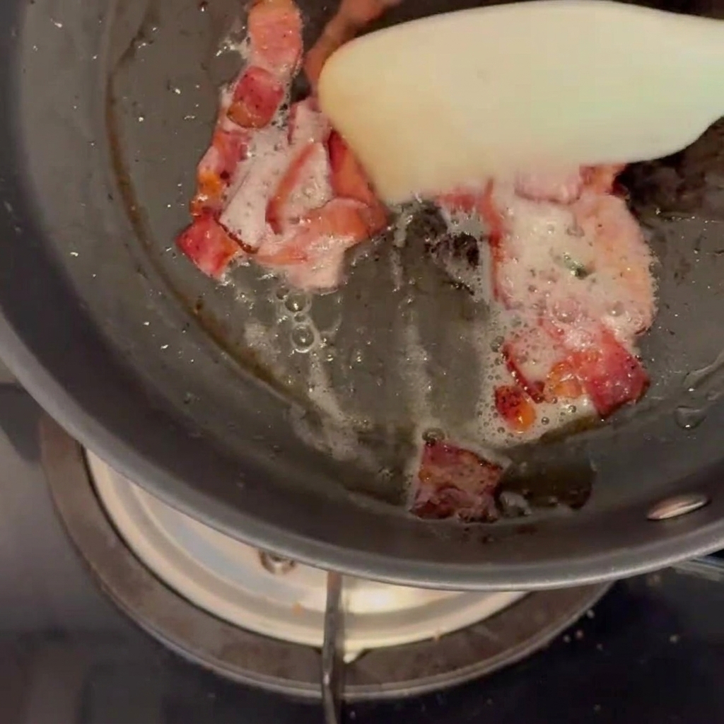 Bacon pieces browning and releasing fat in a frying pan, being stirred with a white silicone spatula.