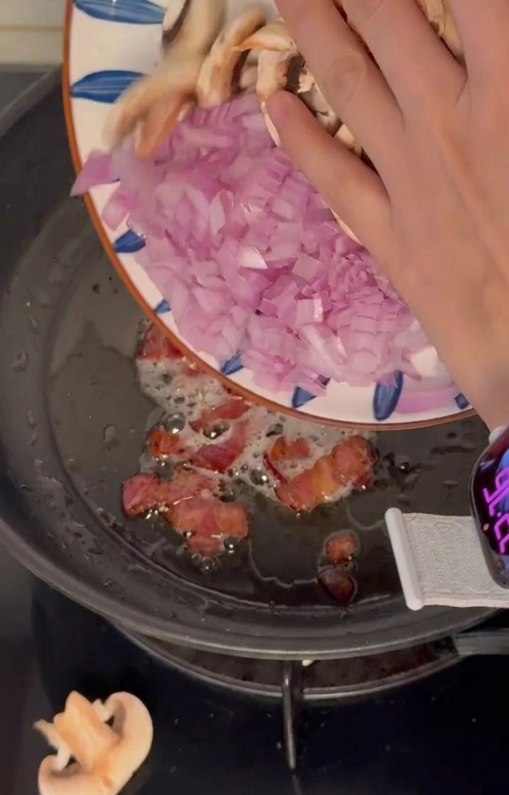 Diced red onions and sliced mushrooms being poured from a plate into a frying pan with bacon.