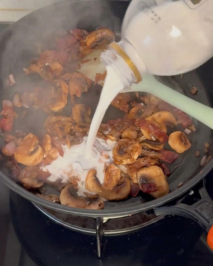 Milk being poured from a plastic bottle into a pan containing cooked mushrooms, onions, and bacon.