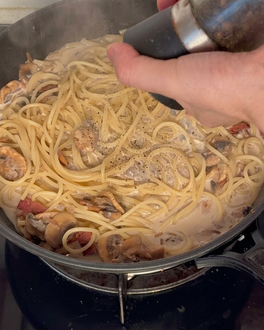 Fresh black pepper being ground over a pan of creamy pasta with mushrooms and bacon.