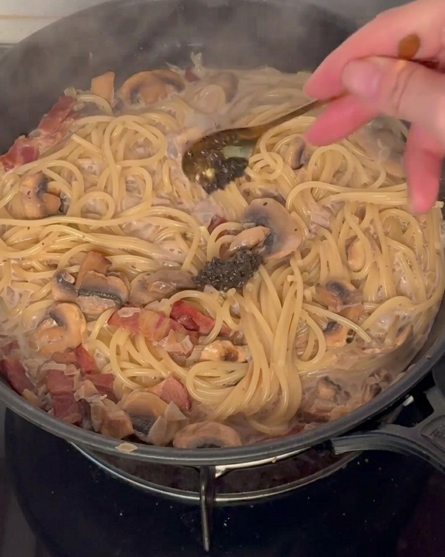 Black truffle paste being added to a pan of creamy pasta with bacon and mushrooms.