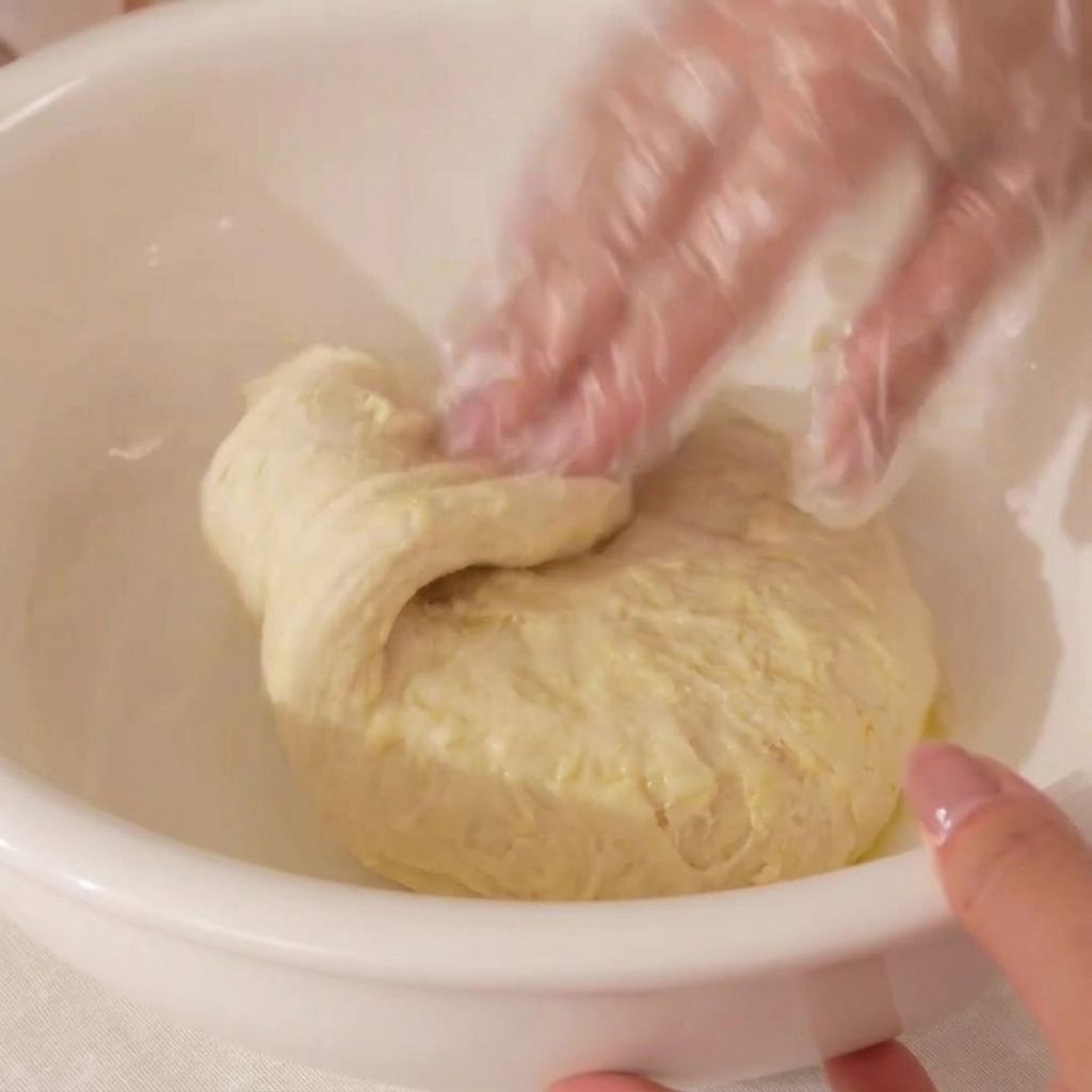 A hand wearing a clear plastic glove mixing pizza dough inside a white bowl.
