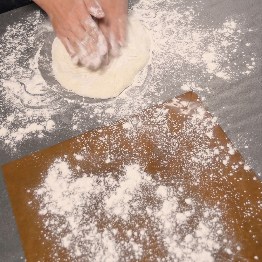 Two hands pressing down on a proofed dough ball on a flour-dusted surface to shape a pizza crust.