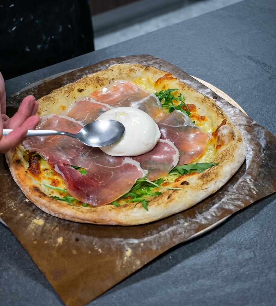A spoon positioning a whole, white burrata cheese ball in the center of a pizza topped with arugula and prosciutto.