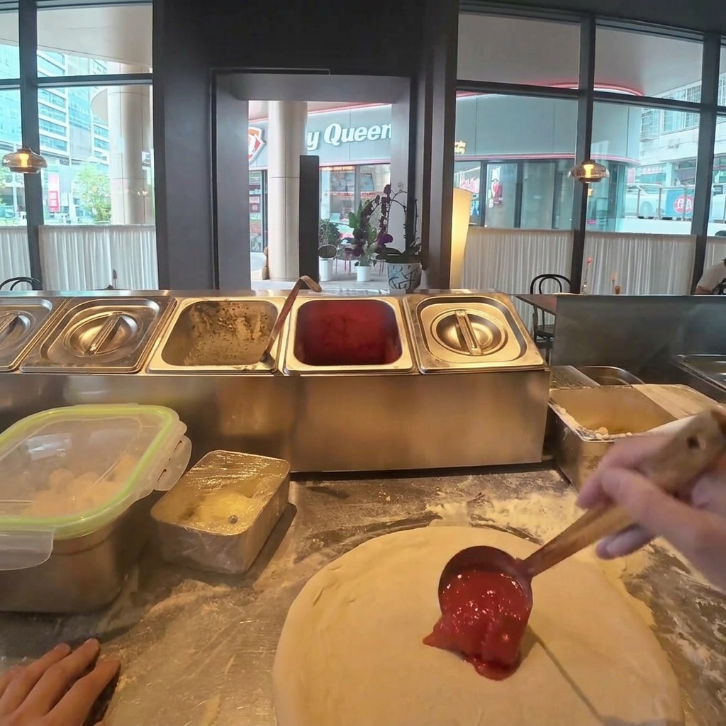 Tomato sauce being spread evenly over the surface of the pizza dough with a metal ladle.