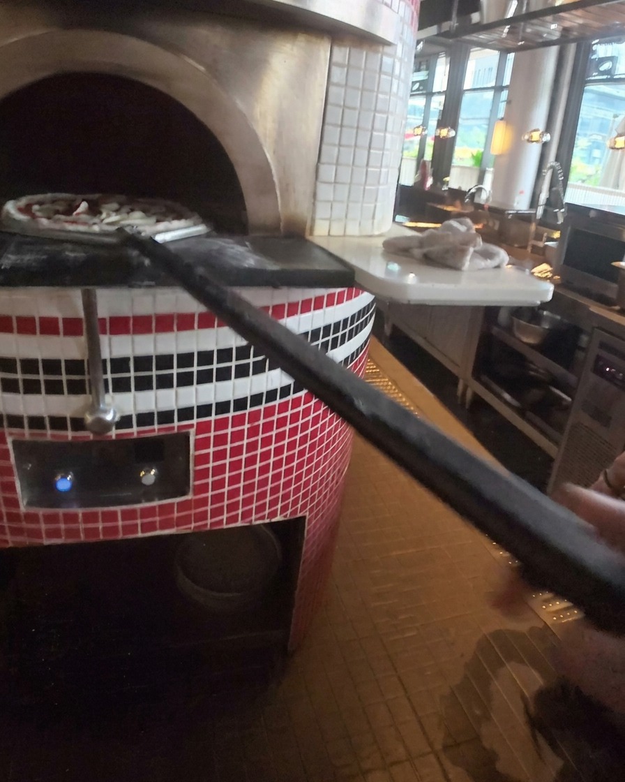 A pizza being slid into a hot stone oven using a long-handled metal pizza peel.