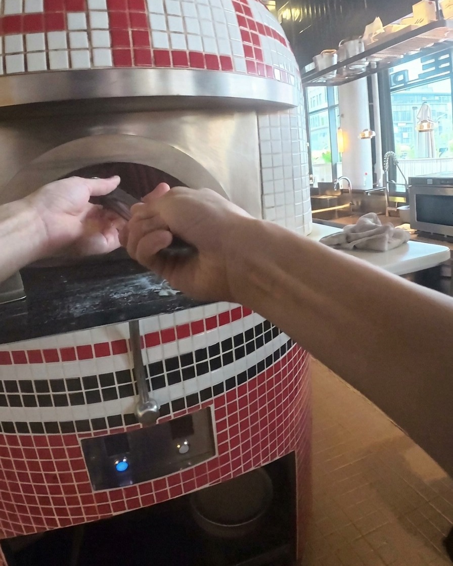 A hot, cooked pizza being lifted out of the stone oven with a metal peel.