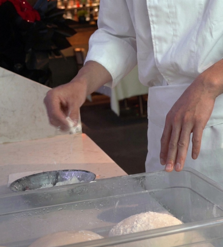 A chefs hands pressing a round ball of pizza dough into a tray of flour.