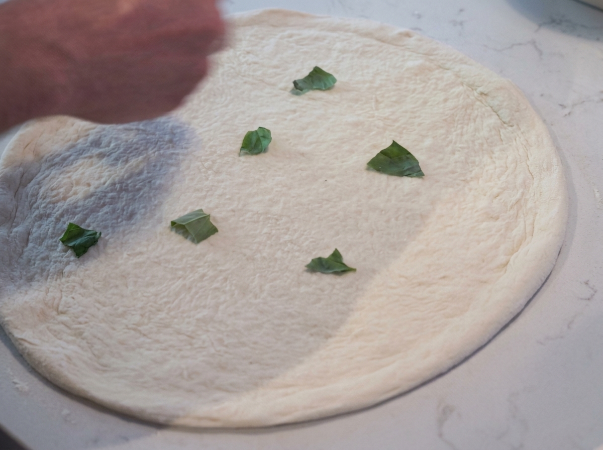 Close-up of fresh green basil leaves scattered on a raw pizza dough base.