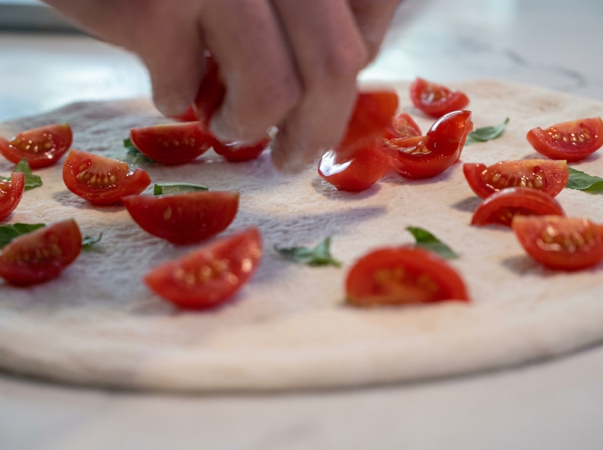 A hand placing halved cherry tomatoes onto a raw pizza dough base garnished with fresh basil leaves.