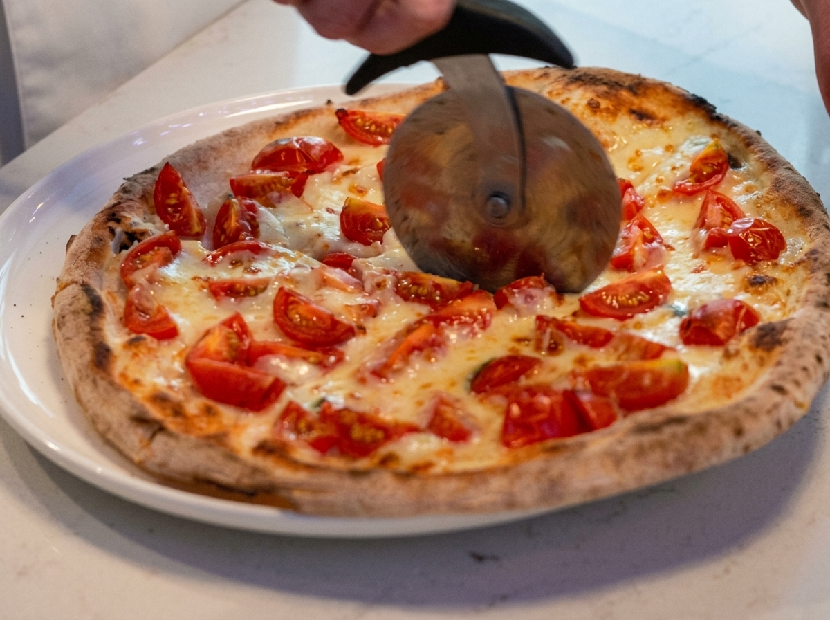 A freshly baked pizza being sliced on a board with a stainless steel pizza cutter.