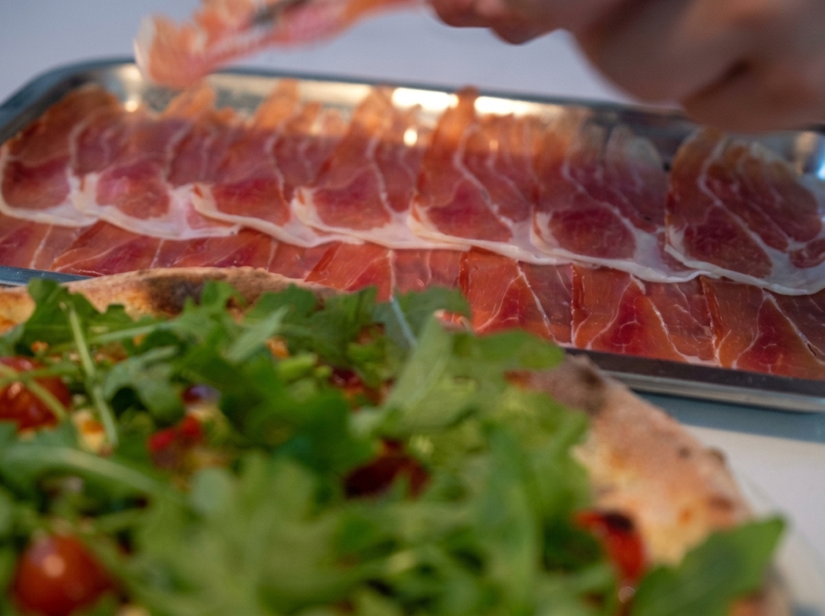 Close-up of thin, delicate slices of Parma ham being carefully placed on top of a bed of fresh arugula on a baked pizza.