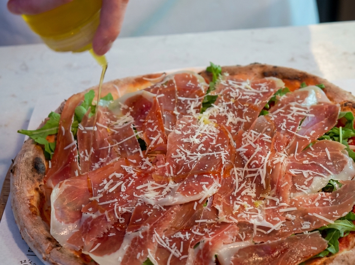 A hand using a squeeze bottle to drizzle olive oil over a pizza topped with arugula, Parma ham, and grated cheese.