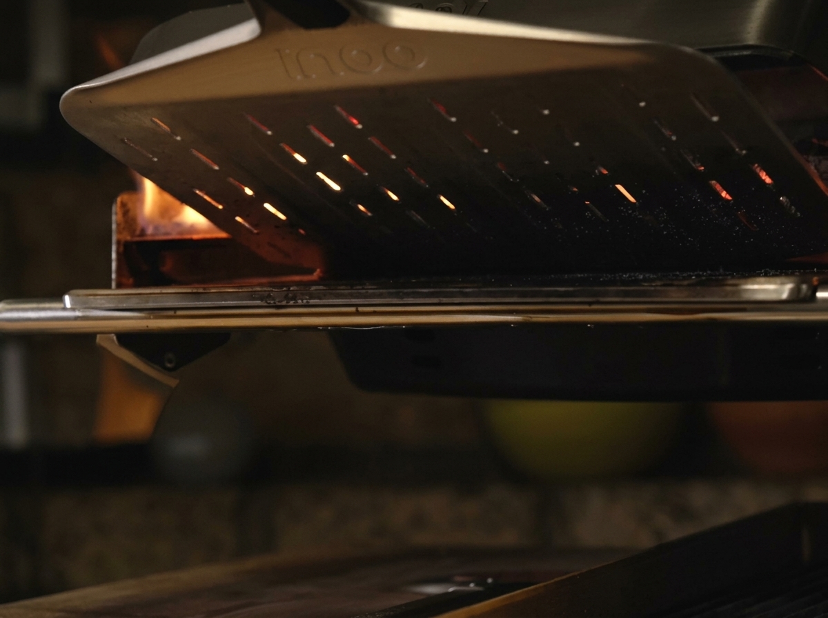 A pizza resting on a metal peel being slid into a glowing, hot portable pizza oven with bright orange flames visible in the back.