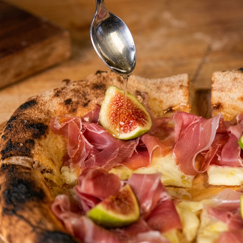 A silver spoon drizzling clear honey over a rustic pizza topped with melted cheese, folded slices of Parma ham, and a halved fresh fig.