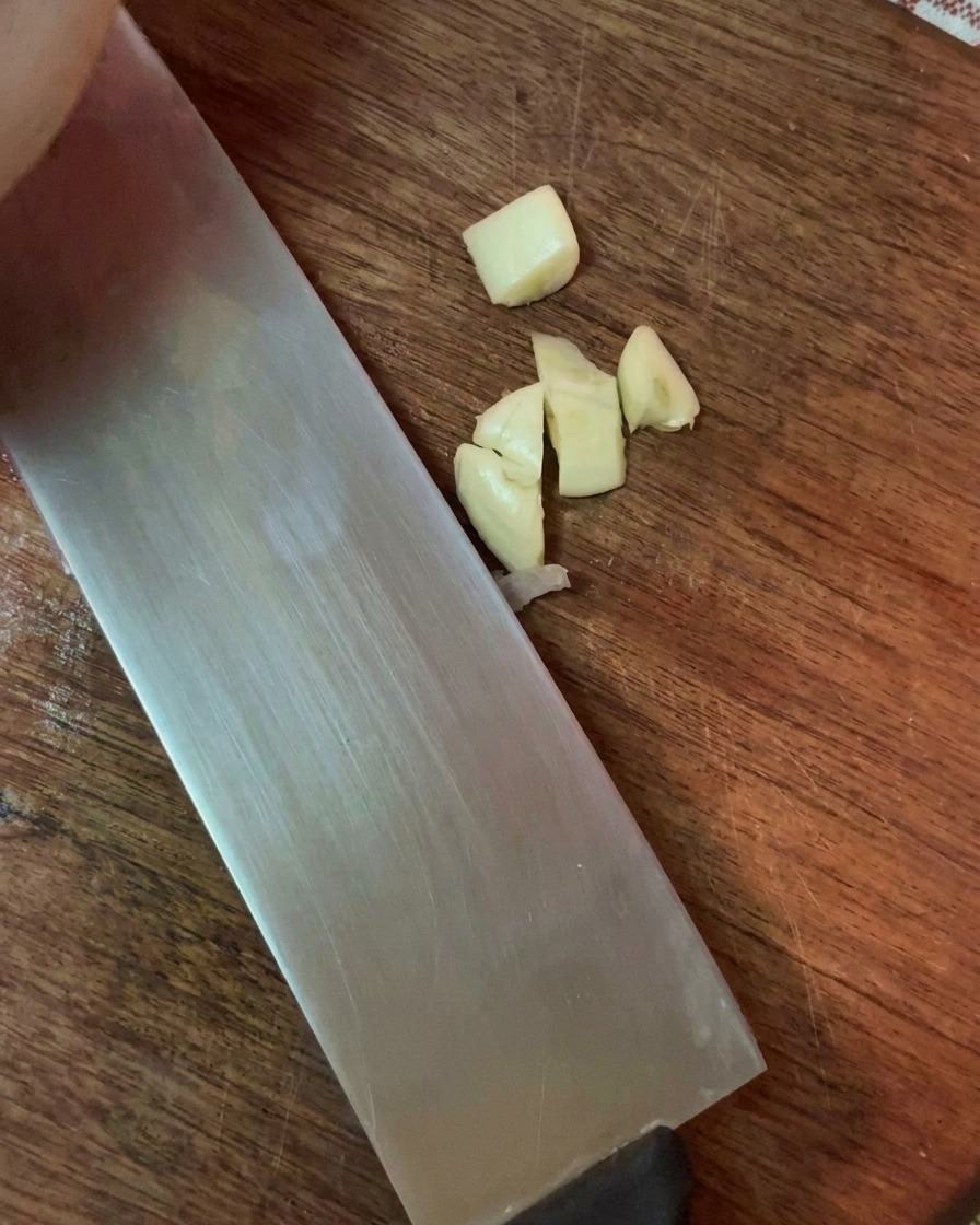 Coarsely minced garlic cloves beside the blade of a wide kitchen knife on a wooden cutting board.