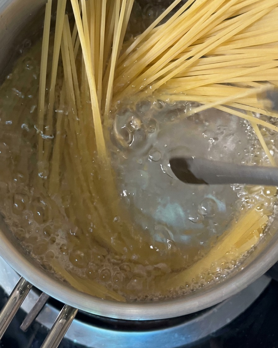 Long strands of dry spaghetti being pushed into bubbling boiling water inside a stainless steel pot.