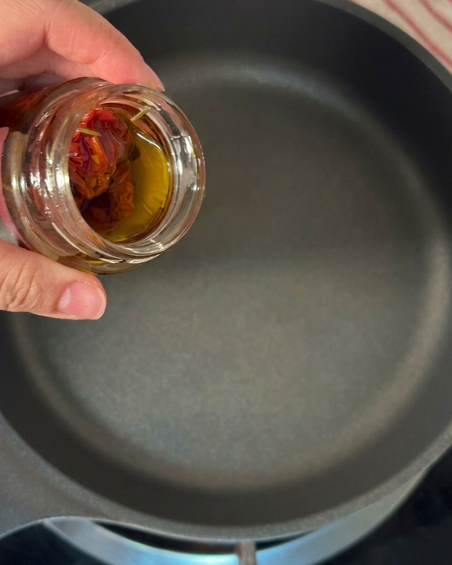 A hand tilting a glass jar of sun-dried tomatoes to pour its golden, herb-infused oil into a large skillet.