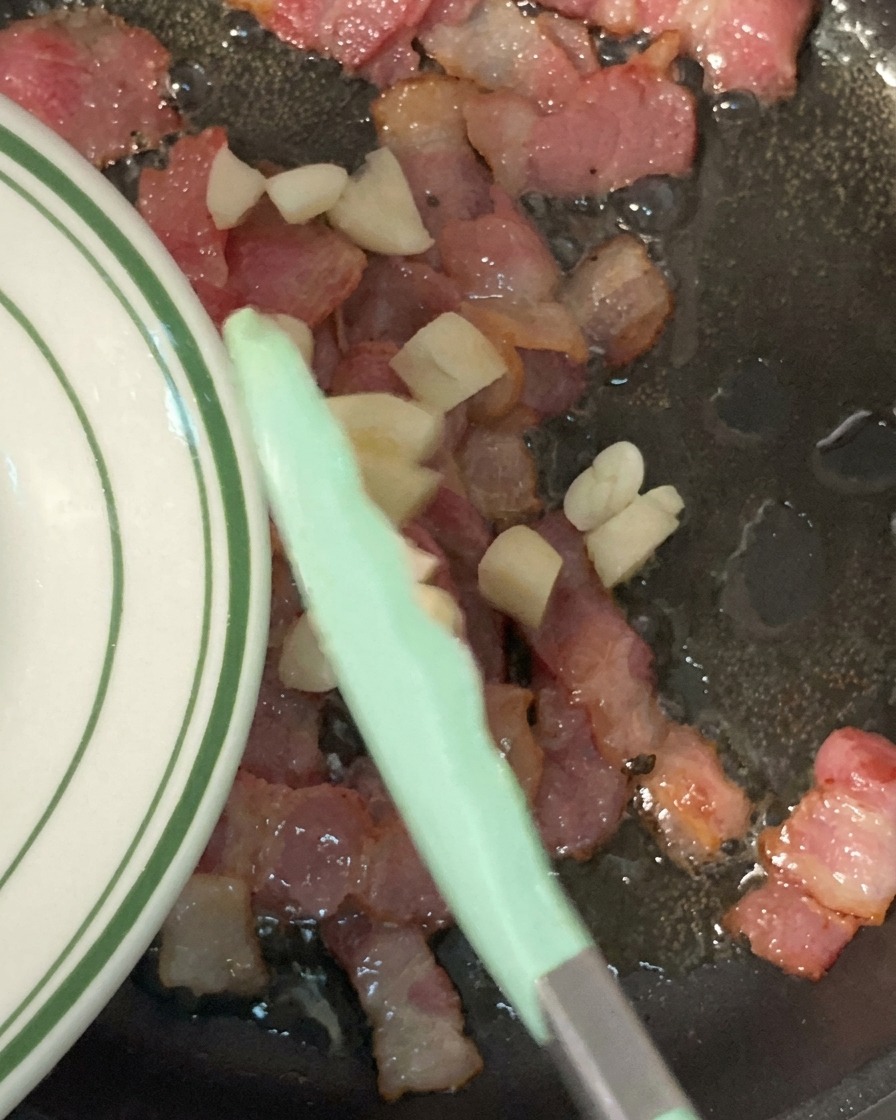 Minced garlic being added from a plate into a skillet filled with chopped, crispy, browned bacon.