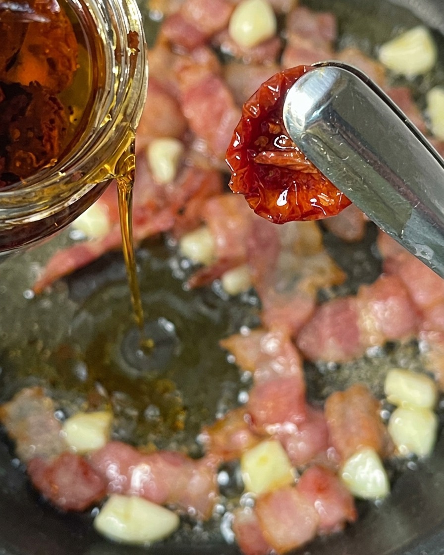 Tongs holding a sun-dried tomato while infused oil is poured from a glass jar into a pan of cooking bacon and garlic.