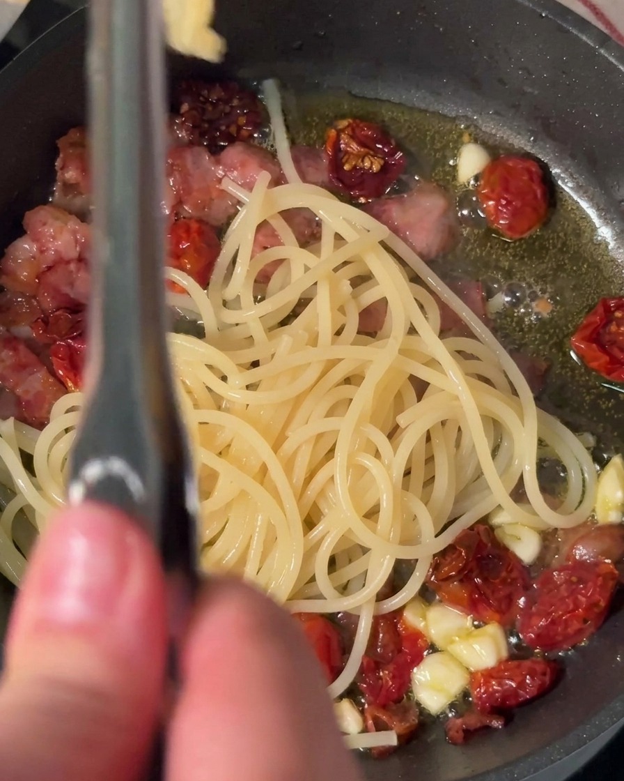 Using tongs to transfer cooked spaghetti noodles directly into a hot skillet filled with bacon, garlic, and sun-dried tomatoes.