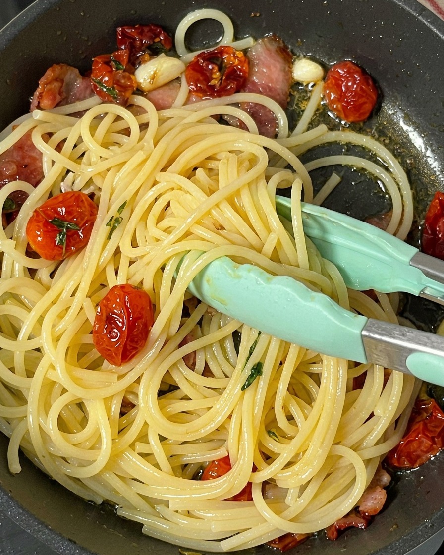 Green silicone-tipped tongs mixing hot spaghetti, bacon strips, and whole sun-dried tomatoes together in a skillet.