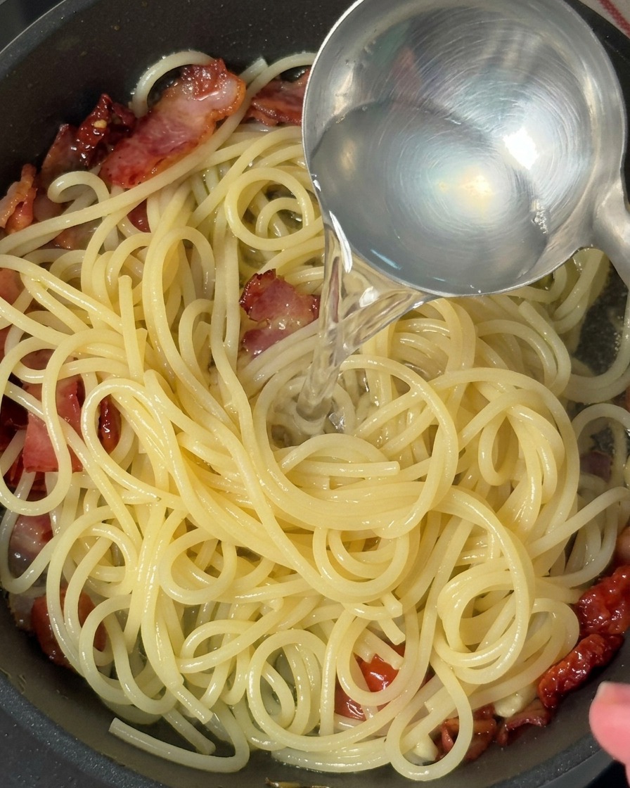 A metal ladle pouring clear, starchy pasta water into a skillet of spaghetti, bacon, and tomatoes.