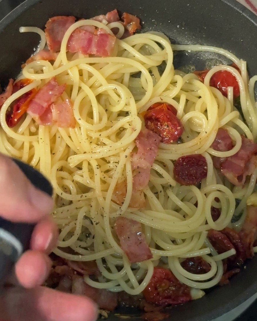 A hand using a black pepper mill to grind fresh black pepper over a pan of cooked spaghetti, bacon, and sun-dried tomatoes.