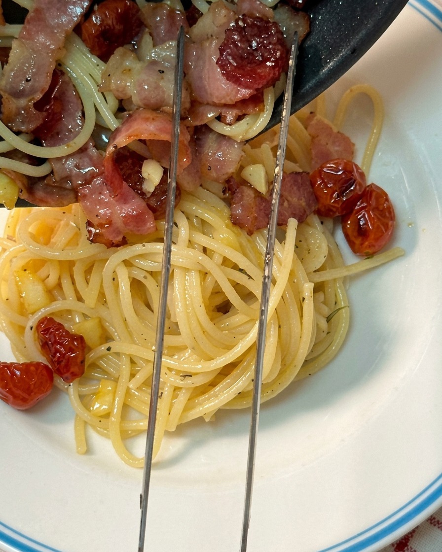 Sliding the finished hot spaghetti, bacon, and sun-dried tomatoes from the black skillet directly onto a white serving plate with a blue rim.