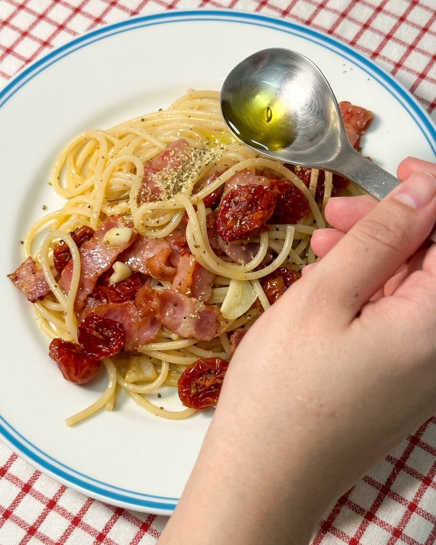 A close-up view of a spoon drizzling golden olive oil over a portion of plated sun-dried tomato and bacon pasta.