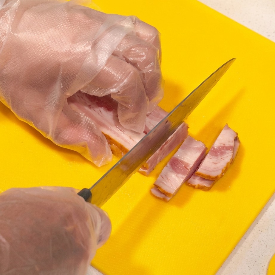 Thick slices of raw bacon being cut into strips on a bright yellow cutting board.