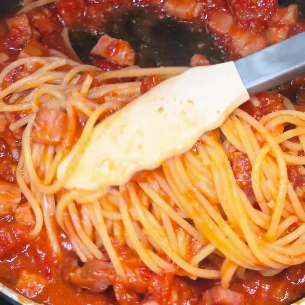 Spaghetti being tossed with tongs in a skillet, becoming well-coated in a vibrant red tomato and bacon sauce.
