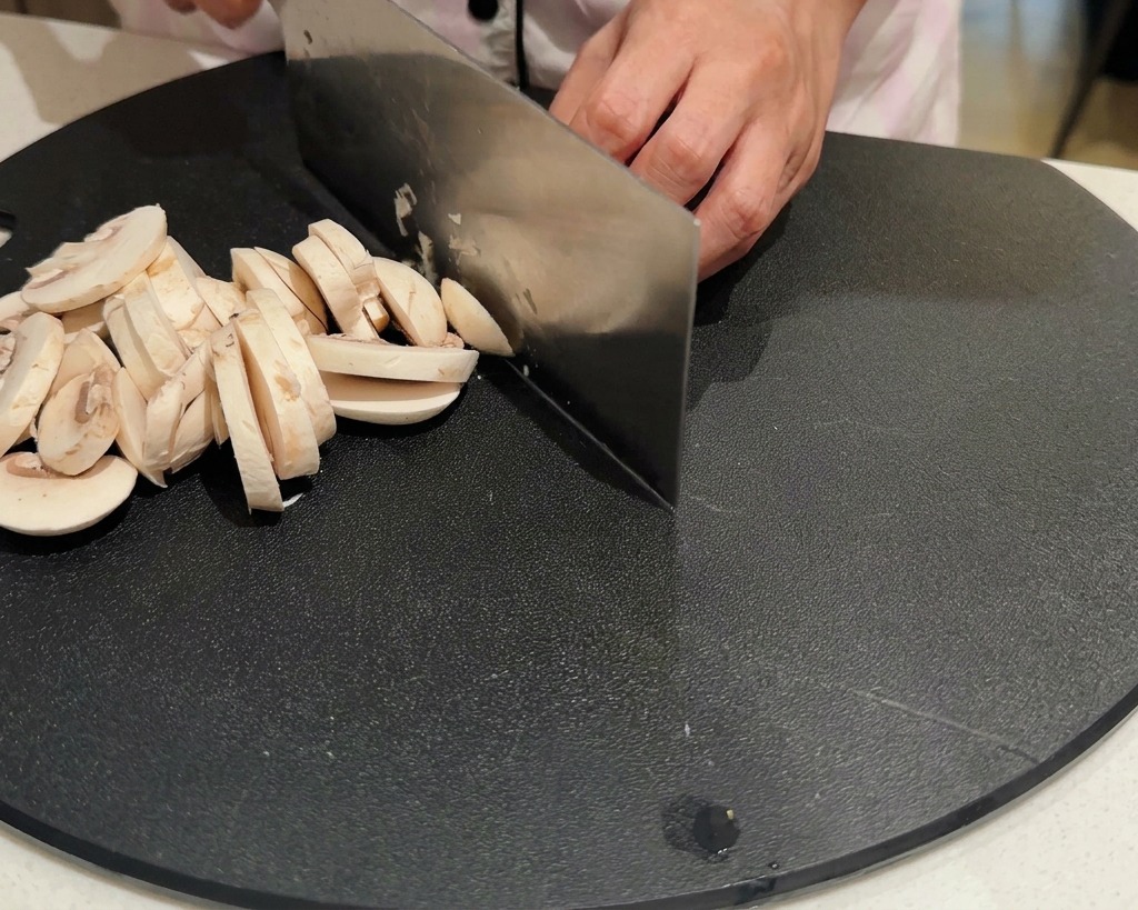 Hands using a large knife to slice fresh white button mushrooms on a black cutting board.