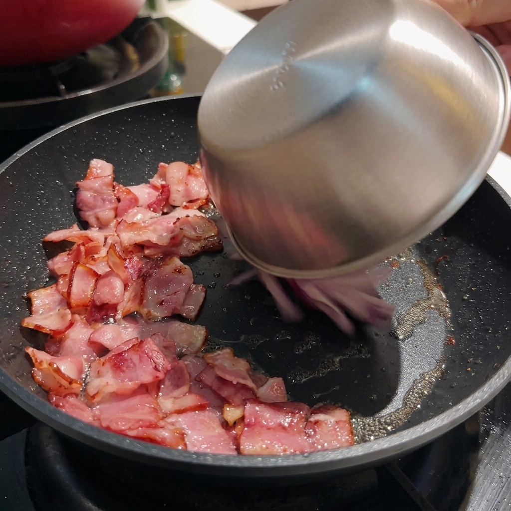 Sliced red onions being poured from a stainless steel bowl into a hot pan containing crisped bacon pieces.