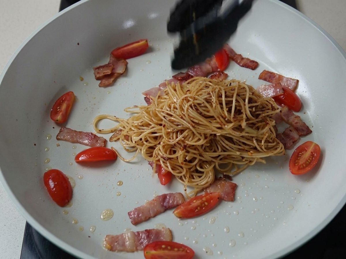 Seasoned spaghetti being tossed with bacon and tomatoes in a pan.